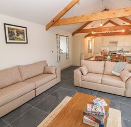 A living room with two beige sofas a wooden coffee table with board games and an open kitchen with wooden beams at The Byre in St Brides Major
