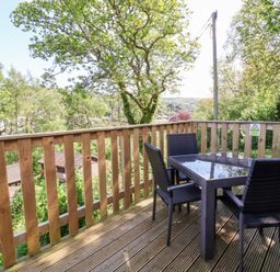 An outdoor wooden deck with a glass-topped table and four black chairs surrounded by trees at Chalet Log Cabin L2 in Combe Martin