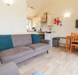 A living area with a sofa two blue cushions a small kitchen with appliances a dining table with chairs and a wall-mounted TV at Chalet Log Cabin C11 in Combe Martin
