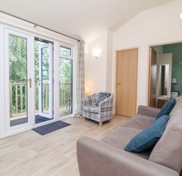 A living room with a sofa with blue cushions an armchair and glass doors opening to a balcony at Chalet Log Cabin C8 in Combe Martin