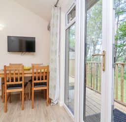 A dining area with a wooden table and four chairs next to a glass door leading to a wooden deck surrounded by trees at Chalet Log Cabin C8 in Combe Martin