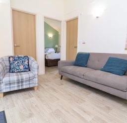 A living room with a plaid armchair and a sofa with blue cushions and open doors showing a bedroom at Chalet Log Cabin C8 in Combe Martin
