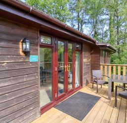 A wooden deck with glass table and three chairs outside a wooden chalet with glass doors at Chalet Log Cabin C9 in Combe Martin