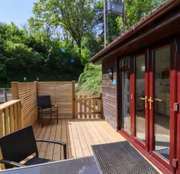 A wooden deck with two black chairs and a table next to a dark wood cabin with glass doors at Chalet Log Cabin C9 in Combe Martin