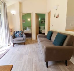 A living room with a sofa and armchair next to open doors showing bedrooms at Chalet Log Cabin L6 in Combe Martin
