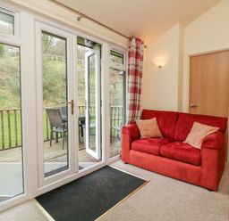 A living room with red sofas beige pillows glass doors opening to a wooden deck with outdoor table and chairs at Chalet Log Cabin L7 Combe Martin