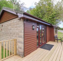 An exterior view of a small building with a wooden deck and two chairs with a table at Chalet Log Cabin L10 in Combe Martin
