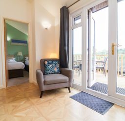 A room with an armchair next to glass doors leading to a balcony with chairs and a view of hills at Chalet Log Cabin L10 in Combe Martin
