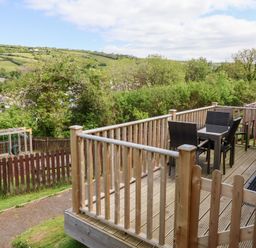 A wooden deck with a table and four chairs overlooking a fenced playground and green hills at Chalet Log Cabin L14 in Combe Martin