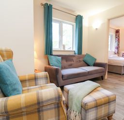 A living room with a plaid armchair and ottoman next to a sofa with teal cushions and a view into a bedroom at Larchwood Lodge in Combe Martin