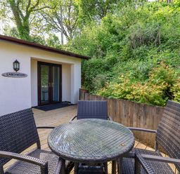 An outdoor wooden deck with a glass top table and four wicker chairs outside a white building with a door and a window at Larchwood Lodge in Combe Martin