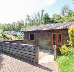 A wooden cabin with a fenced front yard and paved driveway surrounded by trees at Chalet Lodge Bunks L1 in Combe Martin