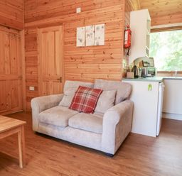 A living area with a beige sofa with cushions a wooden table wooden walls and a small kitchen with a window at Chalet Lodge Bunks L1 in Combe Martin