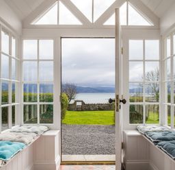 A sunroom with two cushioned benches and glass windows opening to a garden and sea view at Flagstaff in Penmon near Beaumaris