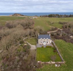 A stone house with a fenced garden surrounded by fields and trees with the sea in the background at Flagstaff Penmon near Beaumaris