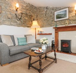 A living room with a gray sofa, wooden coffee table with flowers and plates, a fireplace with a stove and stone walls at Bickle Bice Cottage in Minions near Liskeard