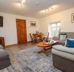A living room with a gray couch a wooden coffee table a round wooden dining table and chairs a wooden door and a wall-mounted TV at Cheesewring Cottage in Minions near Upton Cross