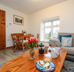 A living room with a wooden table set with scones and coffee mugs a gray couch and wooden dining chairs at Cheesewring Cottage in Minions near Upton Cross