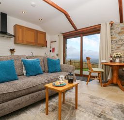 A living room with a brown sofa and blue cushions a small wooden table with a cup and food wooden dining table and chairs near glass doors and a stone wall at Fizz-Gog Cottage Minions near Upton Cross