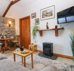 A living room with a wood burning stove small dining table and chairs and a wall mounted television at Fizz-Gog Cottage Minions near Upton Cross