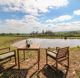 An outdoor seating area with a wooden table and two chairs next to a pond in a rural setting at Tawny in Morchard Bishop