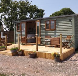 A small wooden cabin with open double doors showing a bed inside a wooden deck with two chairs a table and flower pots at Tawny in Morchard Bishop