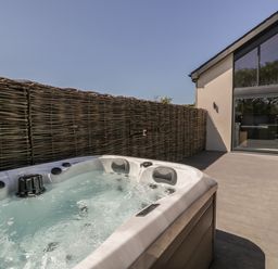 An outdoor patio with a hot tub and a wicker fence next to a modern building with large windows at The Barn in Llanfechell