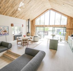 A living room and kitchen with wood-paneled ceiling large windows and grey seating at The Barn in Llanfechell