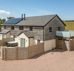 A two-story house with wooden siding and solar panels with a fenced patio and hot tub at Farmyard House in Llanfechell