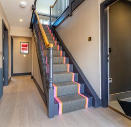 A hallway with stairs covered by a runner including a wooden handrail and two paintings on the wall at Farmyard House in Llanfechell