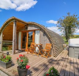 A wooden cabin with curved roof on a deck with chairs flower pots and a hot tub at Pine Lodge in Llanddeusant near Llandovery