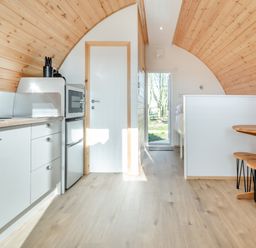 A kitchen area with cabinets microwave and fridge next to a wooden dining table with stools in a pod accommodation at Carlton Manor Pod 2 Aldbrough