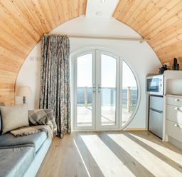A small living area with a couch pillows blanket lamp kitchen appliances and round glass doors leading outside at Carlton Manor Pod 8 Aldbrough