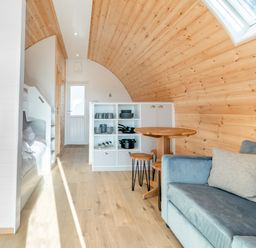 A small living space with wooden walls and ceiling a blue sofa a round wooden table with stools a kitchenette with shelves and kitchenware and bunk beds at Carlton Manor Pod 8 in Aldbrough