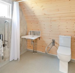 A bathroom with a toilet and sink with support bars and a window with a white curtain at Carlton Manor Pod 10 Aldbrough