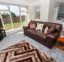A living room with a brown leather sofa and cushions a patterned rug a TV on a stand a wooden side table and a dining area with a white table and chairs at 39 Manorcombe Bungalows in Honicombe Holiday Village near Drakewalls