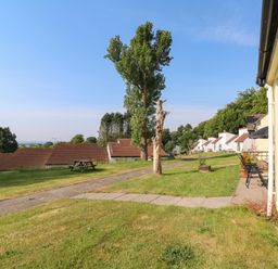 A grassy outdoor area with a paved pathway, trees, chairs, and houses at 39 Manorcombe Bungalows in Honicombe Holiday Village near Drakewalls