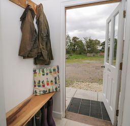 A mudroom with coats hanging on hooks a wooden bench with a cushion and boots underneath an open door showing an outdoor grassy area at Brimley Barn Kittisford near Milverton