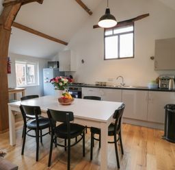 A kitchen with a white table and black chairs a fruit bowl and flowers on the table wooden beams on the ceiling and kitchen cabinets along one wall at Brimley Barn in Kittisford near Milverton