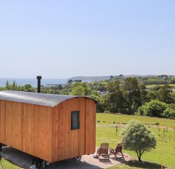 A wooden shepherds hut on wheels near two wooden chairs and a small tree with coastal landscape in the background at Little Pip in Gulval