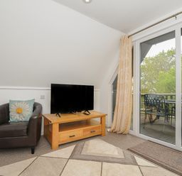 A sitting area with a black armchair with a floral cushion a wooden TV stand with a television and glass doors leading to a patio with outdoor furniture at Valley Lodge 55 in Callington near Gunnislake