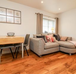 A living room with a grey corner sofa with cushions a small wooden dining table with chairs and a window with curtains at Cosy Cottage in Broadwoodwidger near Lifton