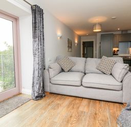 A living room with a grey sofa and patterned cushions near a glass door with grey curtains and an open kitchen in the background at The Old Cowshed Broadwoodwidger near Lifton