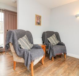 A room with two armchairs covered with gray throws and striped cushions a wooden floor a small wooden table a narrow bench near a door with vertical blinds and a wall light at The Old Cowshed in Broadwoodwidger near Lifton