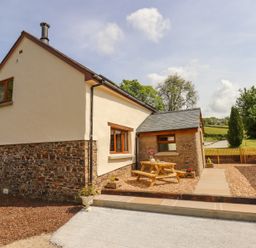 A cottage exterior with a wooden picnic table on a gravel patio and a garden in the background at Bunts Barn Northlew near Hatherleigh