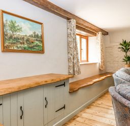 A room with wooden cabinets bench seating by a window patterned curtains a plant and a sectional sofa at Bunts Barn Northlew near Hatherleigh