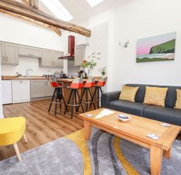 An open plan kitchen and living room with grey cabinets a wooden countertop and a black sofa at Harvenna Cottage in Crackington Haven
