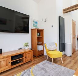 A living room with a yellow chair a wooden TV stand with plants a bookshelf and kitchen area at Harvenna Cottage in Crackington Haven