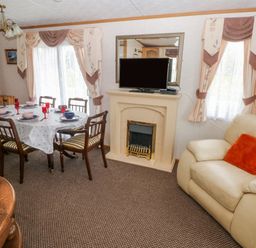 A dining area with a table set for six next to a fireplace with a television and an armchair with an orange cushion at Park Lane Llanteg near Kilgetty