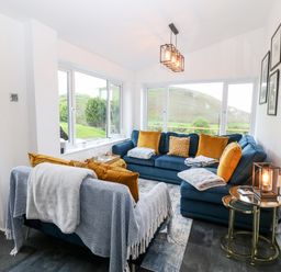 A sitting room with blue sofas orange cushions and blankets with a side table and lamp at Pennant Cottage in Llithfaen
